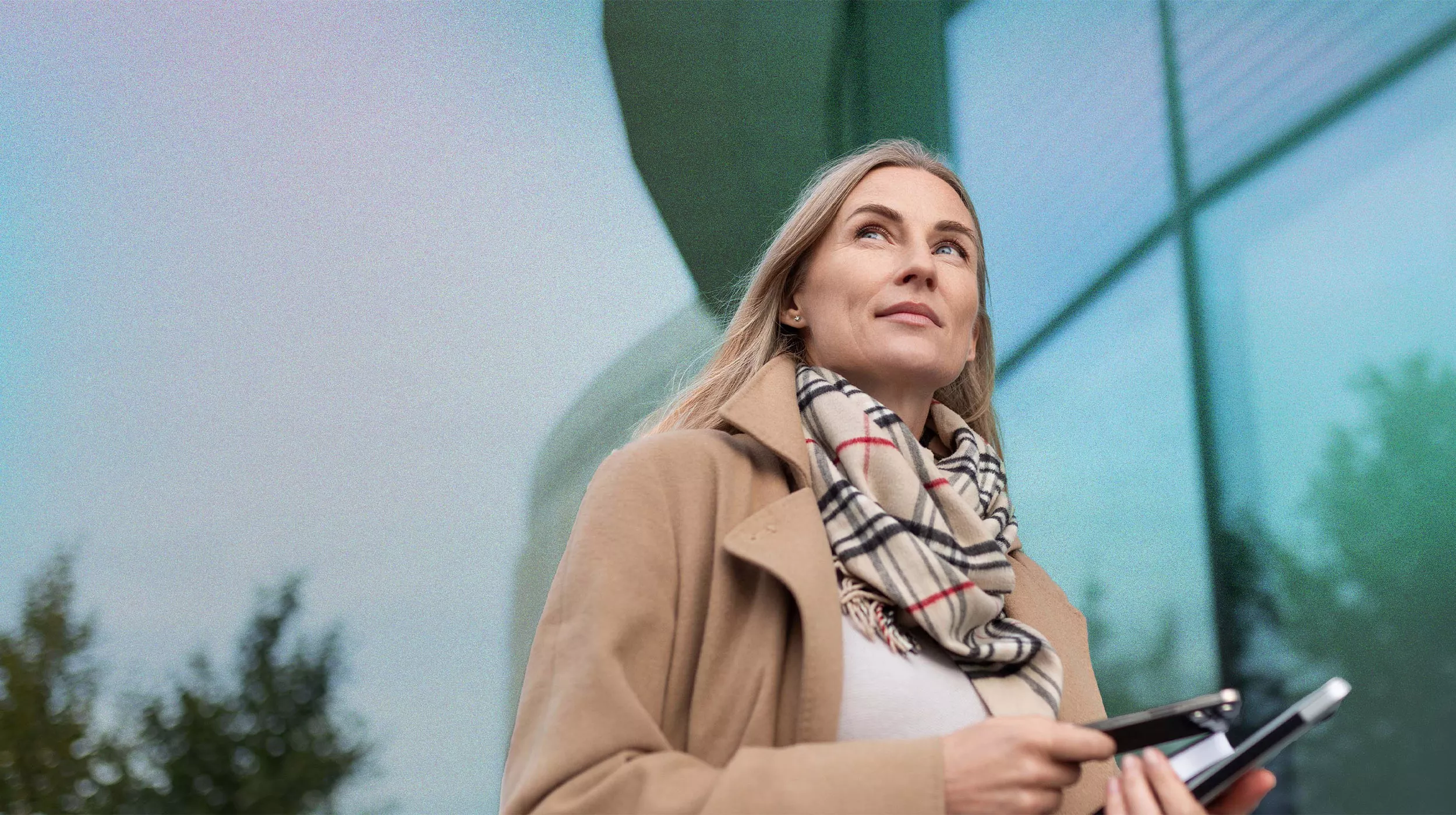 A woman in a beige coat and patterned scarf is holding a smartphone and tablet outdoors. The setting features a modern glass building with reflections and greenery in the background. The scene conveys a professional and urban atmosphere.